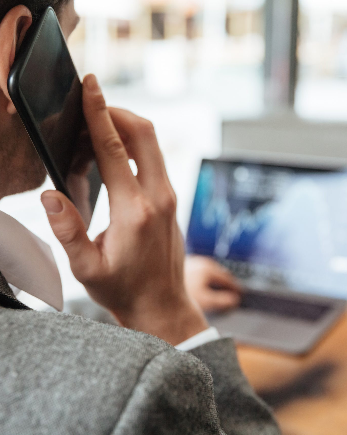 Cropped image of business man sitting by the table in cafe and analyzing indicators on laptop computer while talking by smartphone. Focus on man